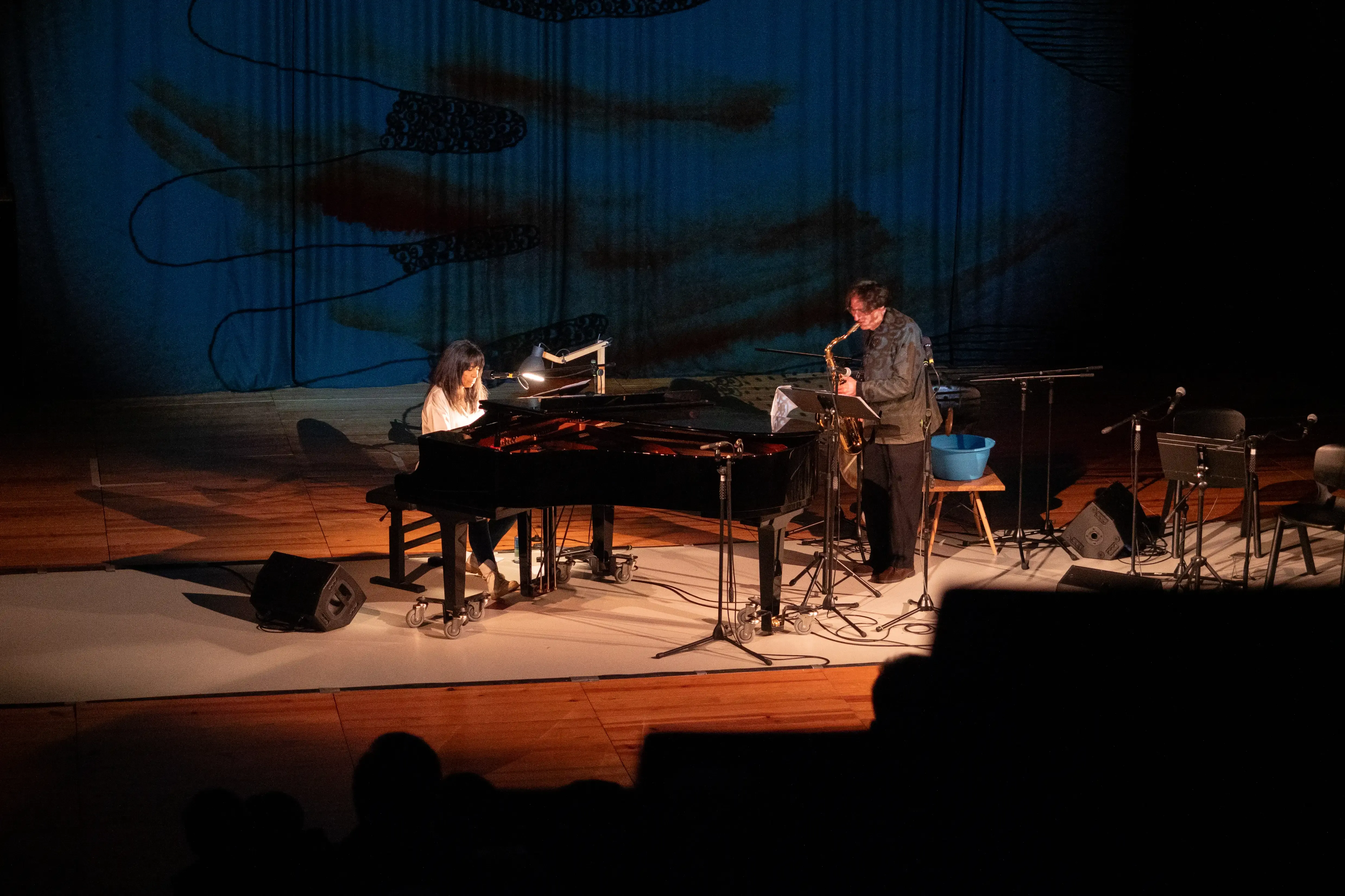 Uma mulher a tocar piano de cauda. Um homem acompanha a mulher com saxofone. No fundo está projetada uma ilustração de uma mão.